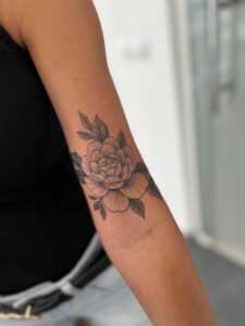 Fotografía de primer plano de un detallado tatuaje de peonía y hojas en tinta negra que cubre completamente el sitio anterior de la cicatriz queloidal en el bíceps superior del brazo.