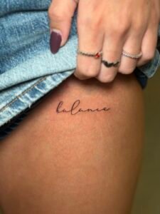 Tatuaje de lettering en letra fina con la palabra balance en la cadera, hecho por Joni Peralta