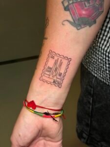 Tatuaje de línea fina de un sello con la Sagrada Familia y BCN en el antebrazo, hecho por Joni Peralta